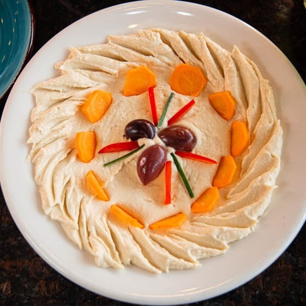 A white plate of hummus garnished with olives, red bell pepper, green onions, and carrots.