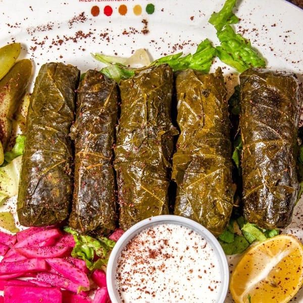 Five stuffed grape leaves (dolmades) with pickles, pink pickled turnips, lemon, and a white dipping sauce.